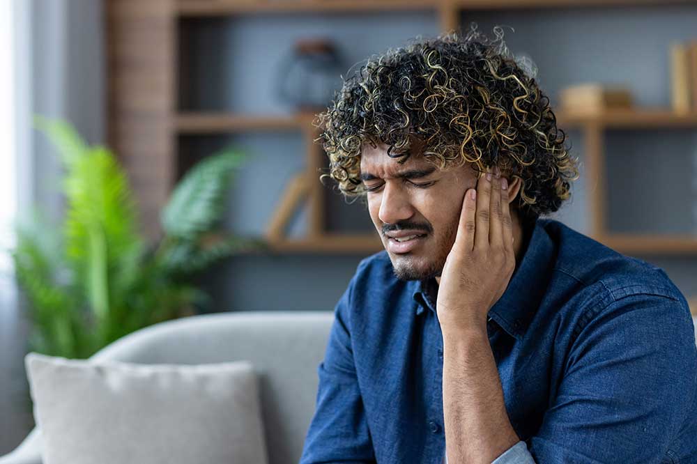 Man with curly hair holding his ear on a couch, bothered by a clogged ear feeling, a concern seen at Audio Recovery in Oklahoma City, OK